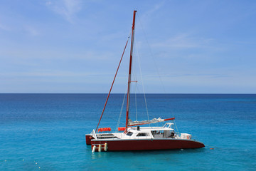 A Catamaran sits at anchor in the bay, Montego Bay, Jamaica. Green and blue water, and a blue sky with light, wispy clouds. Red and White catamaran with sail down.