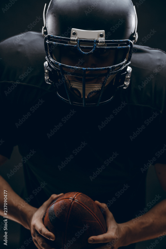 Wall mural American football player with black helmet and armour running in motion, holding ball, getting ready to score a goal, close up shot over dark background