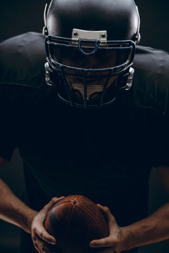 American football player with black helmet and armour running in motion, holding ball, getting ready to score a goal, close up shot over dark background
