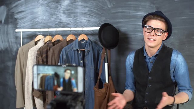Medium shot of young man in fedora felt hat and glasses sitting against chalkboard background and talking about fashion when recording video blog on smartphone
