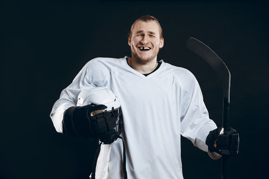 Happy Handsome hockey player with one broken front tooth laughing at camera, standing with stick in white uniform, isolated on black