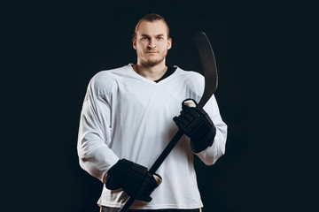 Fototapeta premium Portrait of confident professional hockey in white uniform player holds gaming stick isolated on black background.