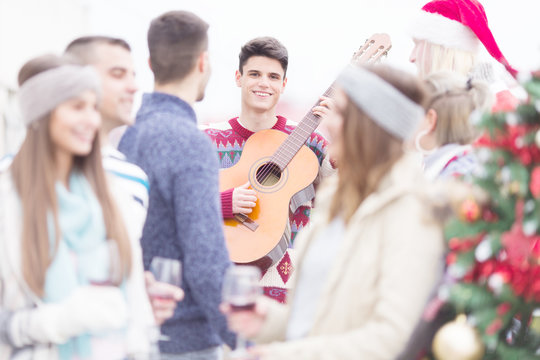Young man with guitar smiling and celebrating Christmas on balcony with friends - Powered by Adobe