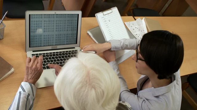 High angle view of young woman holding clipboard with document attached to it and taking notes while elderly woman sitting next to her and taking online test on laptop computer