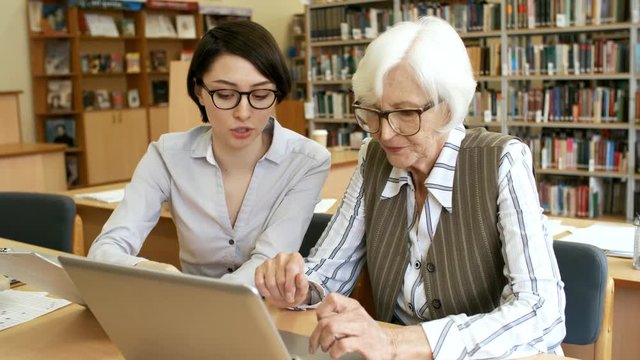 Medium shot of young woman in glasses sitting next to senior lady and helping her learn to use laptop computer