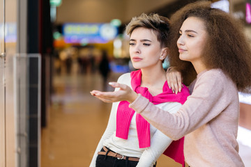 Two delighted diverse young women being in great mood while doing shopping in a big city mall. Young blonde woman showing something in the shop showcase to her female friend with brown frizzy hair