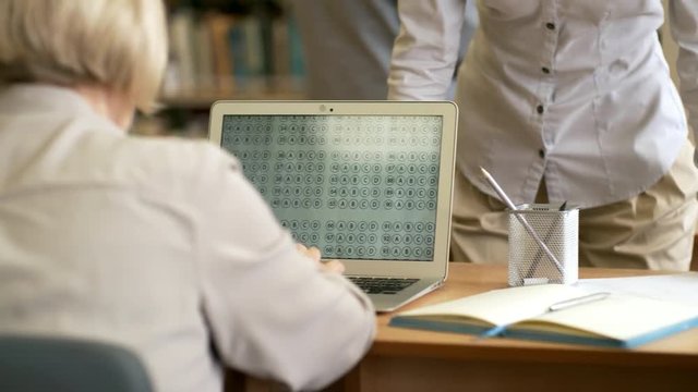 Rear view medium shot of group of retired people taking online test using laptop computers, unrecognizable female teacher explaining task to them, tracking left