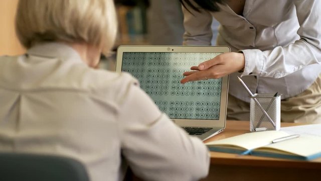 Rear view medium shot of retired woman using laptop computer when taking online test, unrecognizable female teacher explaining task to her