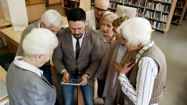 High angle view of group of retired people standing around middle-aged man with tablet computer and talking to him