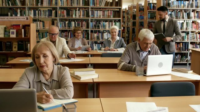 Group of concentrated retired people sitting at desks in library and studying, male teacher monitoring their progress and helping them