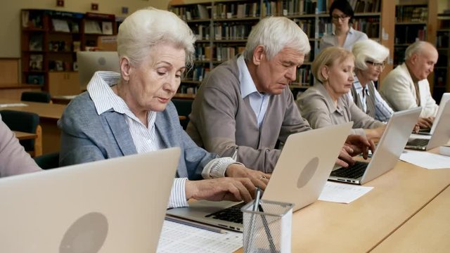 Group of elderly people sitting next to each other in line at desks in library and using laptop computers while young female teacher speaking to them during computer class