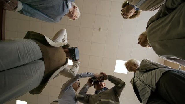 Low angle view of group of retired people standing in circle and listening to young female teacher of information technology, two of them trying virtual reality goggles