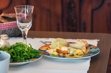 A plate with salmon, potatoes, carrot and cream sauce with lemon on a wooden table with a water glass, home background.