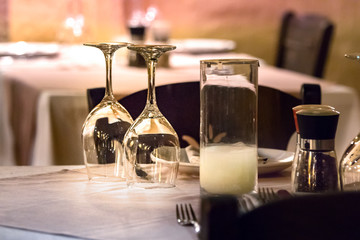 Two wine glasses upside down on a table with shakers, plates and cutlery at evening, outdoors.