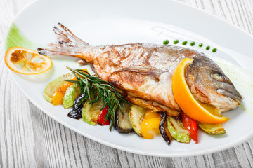 Grilled dorado fish with baked vegetables and rosemary on plate on wooden background close up. Healthy food. Top view