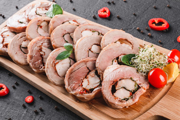 Assorted meat, stuffed chicken roll, meat roll with pepper, greens on cutting board on black shale background. Meat appetizer, food concept. Top view, flat lay
