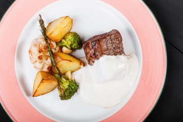 Roasted potatoes with beef meat, herbs, broccoli and sauce in plate on dark wooden background. Hot Meat Dishes. Top view, flat lay