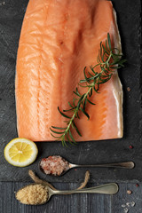 Raw salmon fillet on black slate plate and ingredients for making gravlax. Salt, sugar, lemon and rosemary in the background for dry cure marinade, dark background.