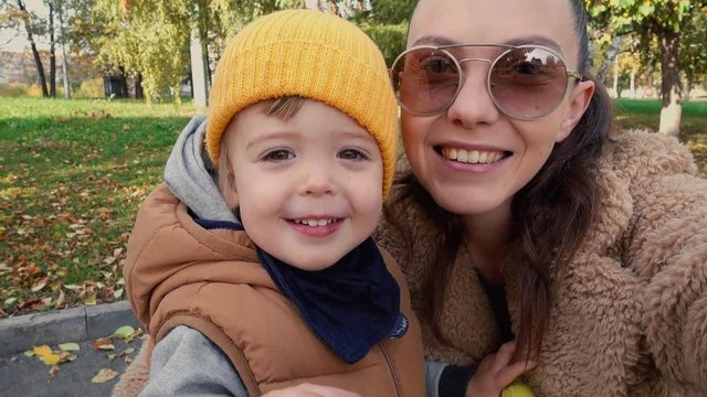 Stylish young woman in sunglasses taking selfie with little son waving with hand at camera in park
