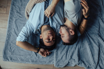 Happy homosexual couple under the white blanket on the bed. Guys lie on back, both smiling feeling tenderness and love. Closeup. Top view.