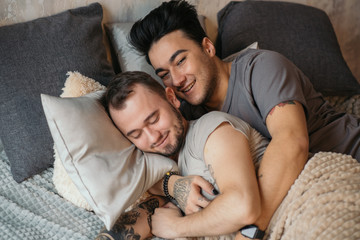 Close up shot of appealing happy gay couple smiling while resting in bed and dominant guy embracing...
