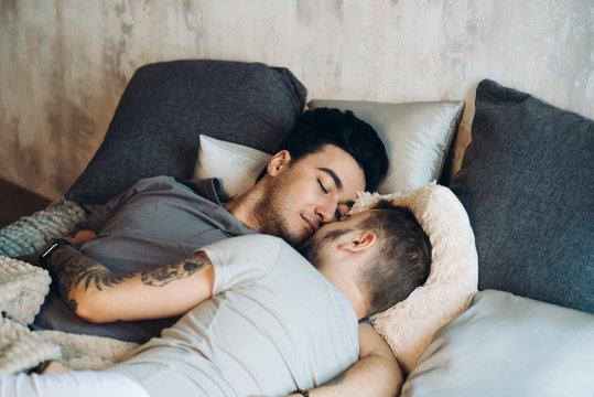 Caucasian Homosexual Family In Bed Before Sleep. Handsome Hot Man With Bristle Kissing Good Night His Sleepy Male Partner, Lying In Bed