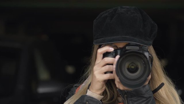Professional young blonde woman photographer with digital camera taking pictures at city parking. Close Up, shallow depth of field, dark background.