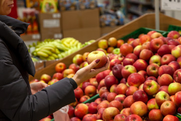 A young blonde woman in a long jacket chooses food while shopping in a mall in the fruit department holding an apple of red-green color in her hand among a lot of ripe products.