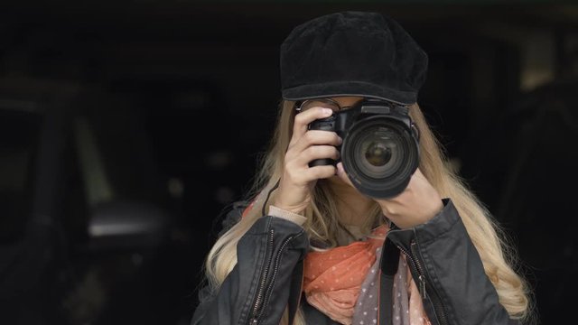 Professional young blonde woman photographer with digital camera taking pictures at city parking. Close Up, shallow depth of field, dark background.