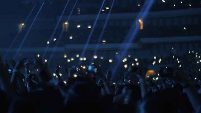 Wide shot of hands moving in time with the music among cheering fans at a live music concert with blue lights