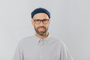 Horizontal view of pleased smiling man wears glasses, hat and shirt, being satisfied with good news, models against white background. Unshaven hipster rejoices new purchase. Fashion concept.