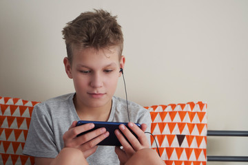 Teen boy watching something on a smartphone  with earphones at home. Teenager watching video on...