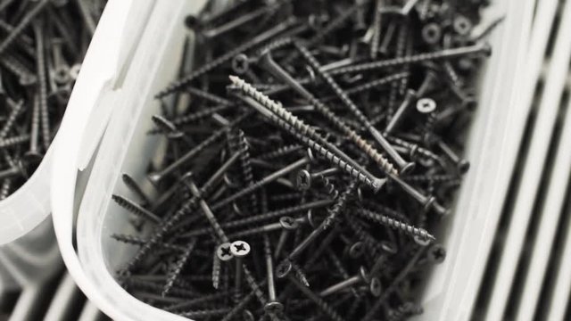 Camera is showing big white plastic containers which is full of long screws and nails placed somewhere in workhouse room.