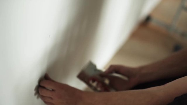 Man's hands are professionally and actively driving heavy staples into white wallpapers by using staple-gun somewhere in workhouse room.
