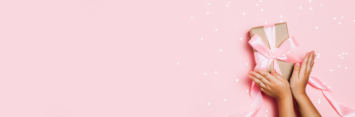 Child hands holding beautiful gift box on pink background, with sparkling glitter around. Holiday, bright and festive view, flat lay. Copy space for text. Christmas greeting banner.