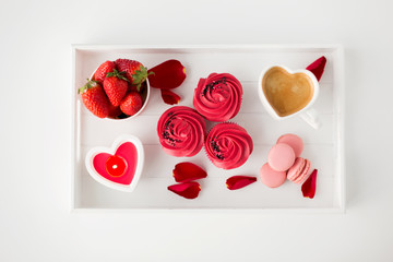 valentines day and sweets concept - close up of cupcakes with red buttercream frosting, macarons, heart shaped coffee cup, candle and strawberries on tray with rose petals on white background