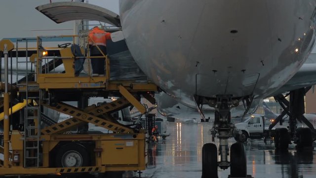 A rainy and busy evening at the airport courtyard. An airport worker is standing on a yellow elevator close to the plane. A massive load is moving out of it. There are some cars on the background, a