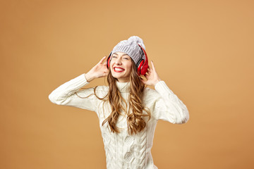 Glad girl in red headphones dressed in white knitted sweater and hat is having fun on a beige background in the studio