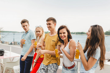 A company of good-looking friends laughing and drinking yellow cocktails in the nice cafe next to the river. Cheers. Entertainment, having good time. River is in the background.