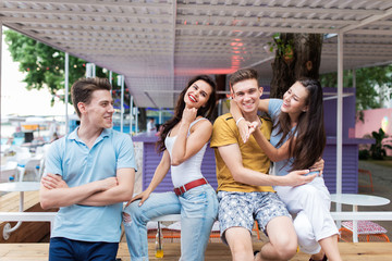 A company of good-looking friends is laughing and sitting on the railing in the nice summer cafe. Entertainment, having good time. Friendship.
