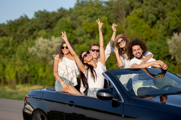 Cool stylish young dark-haired girls and guys in sunglasses smile in a black cabriolet on the road...