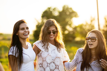 Three beautiful dark-haired young women in sunglasses dressed in the beautiful clothes are smiling and walking outdoor on a sunny day.