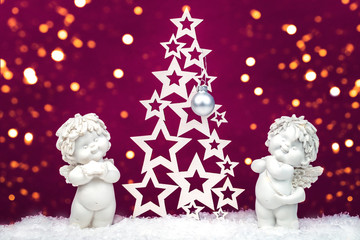 Two sweet Christmas cherub baby angels statuettes on snow with wooden Christmas tree on background with golden glowing lights, New Year and Christmas festive concept