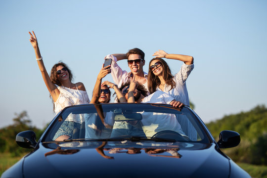 Funny young girls and guys in sunglasses are sitting in a black cabriolet on the road holding their hands up and making selfie on a sunny day.