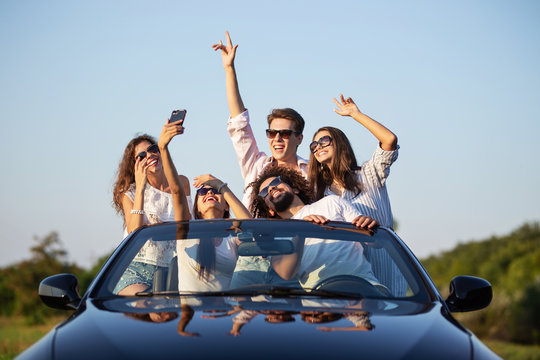 Funny young girls and guys in sunglasses are sitting in a black cabriolet on the road holding their hands up and making selfie on a sunny day.