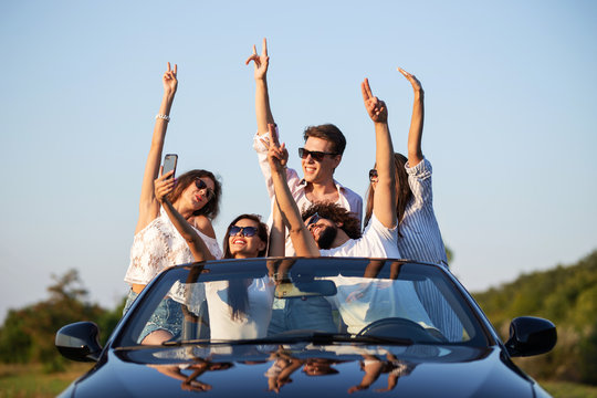Young lucky girls and boys in sunglasses are sitting in a black cabriolet on the road holding their hands up and making selfie on a sunny day.
