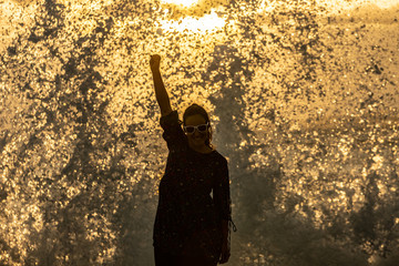 Woman celebrating with clenched fist and breaking big wave behind with sunset on the beach. Powerful girl with her arm and sunglasses on the rough sea