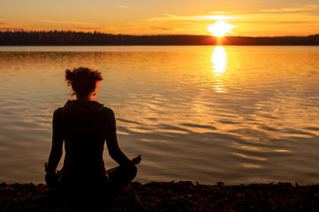 Silhouette of young woman practising yoga outdoor. Meditation on the shore of the lake during sunset. Fitness and healthy lifestyle concept.