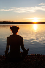 Atmospheric shot of young woman practising yoga in nature. Meditation on the shore of the lake during sunset.  Sun is setting behind the forest trees.