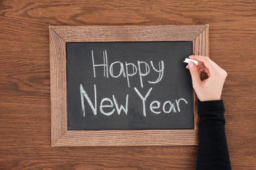 partial view of woman writing 'happy new year' text on chalk board with wooden background